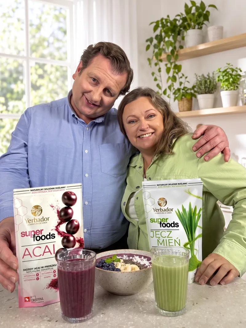 Uśmiechnięta para w jasnej kuchni: mężczyzna w błękitnej koszuli obejmuje kobietę w zielonej koszuli, przed nimi Yerbador Superfoods Açaí i Jęczmień, fioletowy i zielony koktajl oraz bowl açaí