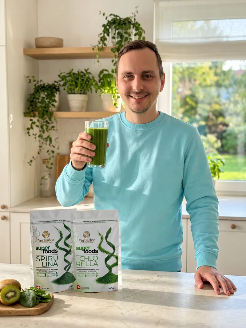Uśmiechnięty mężczyzna w turkusowej bluzie unosi szklankę zielonego smoothie, na blacie Yerbador Superfoods Spirulina i Chlorella, deska z kiwi i liśćmi szpinaku