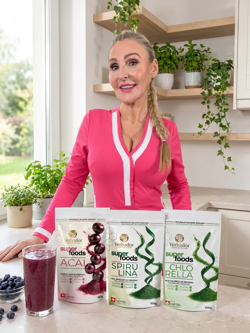 Blondynka z długim warkoczem w różowej sukience stoi przy kuchennym blacie, przed nią Yerbador Superfoods Açaí, Spirulina i Chlorella, szklanka koktajlu açaí i miseczka borówek