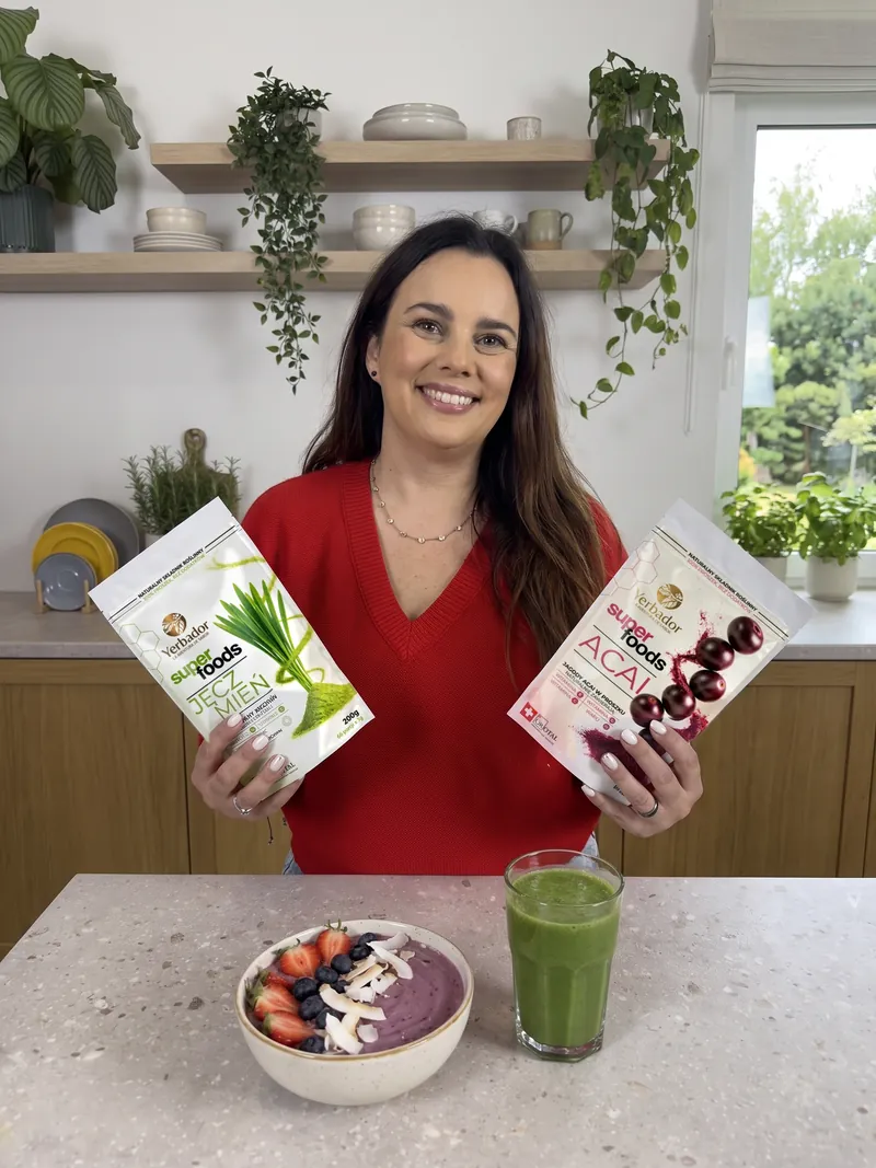 Uśmiechnięta brunetka w czerwonym swetrze trzyma w dłoniach Yerbador Superfoods Jęczmień i Açaí, na blacie bowl açaí z truskawkami i szklanka zielonego smoothie