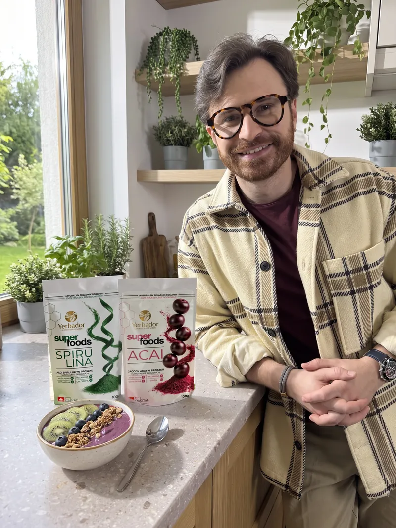 Mężczyzna z brodą, w okularach i kremowej koszuli w kratę opiera się o kuchenny blat, obok Yerbador Superfoods Spirulina i Açaí oraz bowl açaí z kiwi, borówkami i granolą
