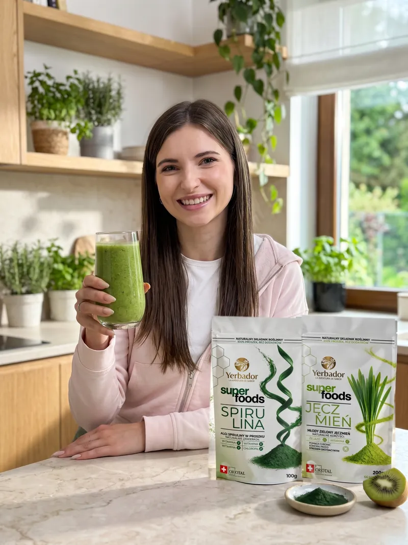 Uśmiechnięta brunetka z długimi prostymi włosami w jasnoróżowej bluzie unosi szklankę zielonego smoothie, na blacie Yerbador Superfoods Spirulina i Jęczmień, miseczka zielonego proszku i połówka kiwi