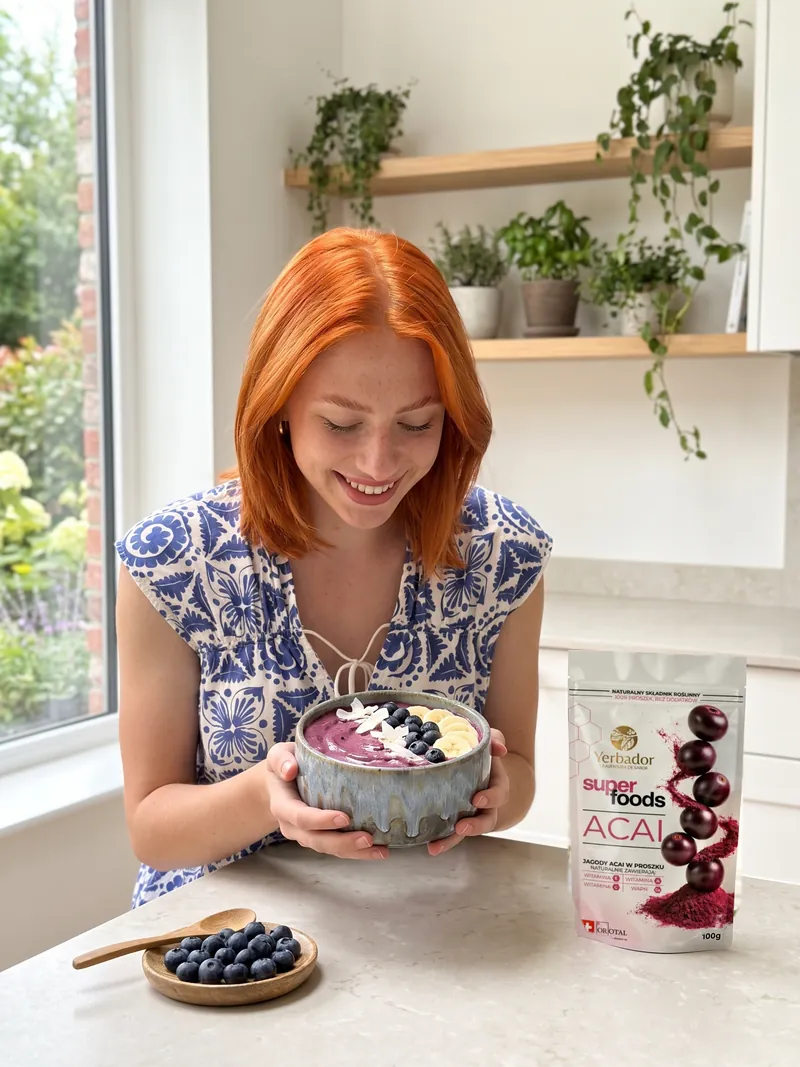 Rudowłosa kobieta w biało-niebieskiej wzorzystej bluzce trzyma w dłoniach ceramiczną czarkę z bowlem açaí z borówkami, bananem i kokosem, obok Yerbador Superfoods Açaí i miseczka borówek