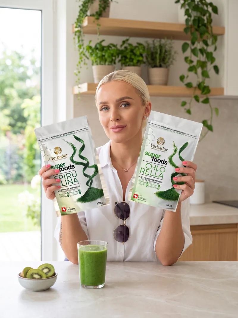 Blondynka z gładko zaczesanymi włosami w białej koszuli trzyma w uniesionych dłoniach Yerbador Superfoods Spirulina i Chlorella, na blacie szklanka zielonego smoothie i miseczka kiwi