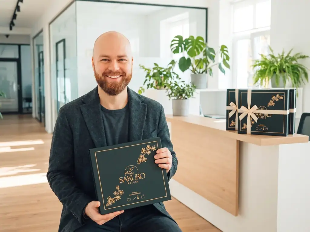 Yerba mate gabinet kosmetyczny: uśmiechnięty mężczyzna z brodą trzyma pudełko prezentowe Sakuro Matcha na recepcji biura
